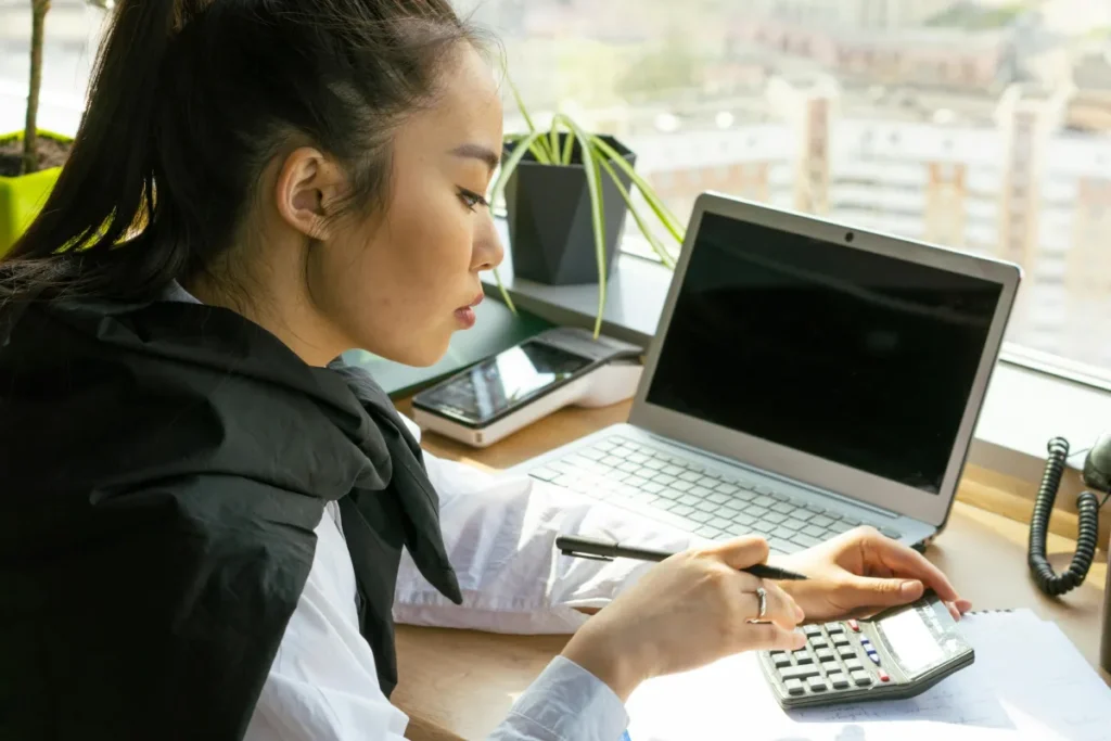 Perempuan bekerja menggunakan laptop dan kalkulator di meja kantor terang untuk mengelola keuangan bisnis